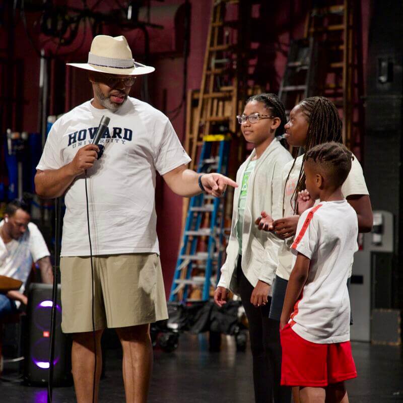 Eric Roberson in tan shorts, white shirt and straw hat speaking with students