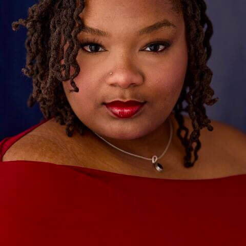 Sunday Coleman, African American female with brown loc'd hair that drapes alongside her face, with a nose piercing, silver necklace, wearing an off the shoulder burgundy shirt
