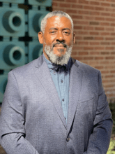 Black man with gray and black hair and beard, blue shirt and suit jacket