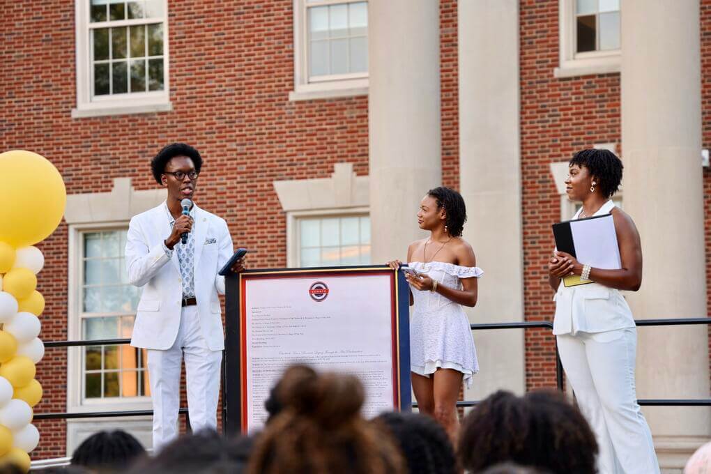 Fine Arts Student Council President Noel Best (right) looks on as Chris Moore III (left) and [NAME] read the Chadwick Aaron Boseman Legacy Through the Arts Proclamation to the crowd during the Student Showcase.