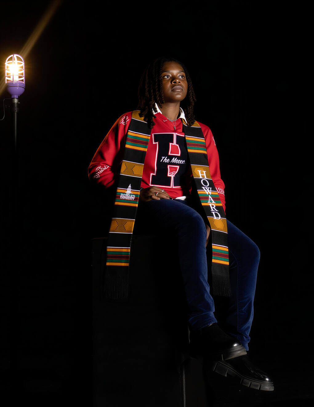 Jaadyn Rogers poses in Howard University apparel and a Kente stole, illuminated against a dramatic black backdrop.